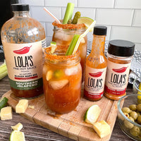 A group of bottles of Lola's Southern Jalapeño Bloody Mary Mix on a cutting board, with a jar of hot sauce, citrus fruits, and green olives.