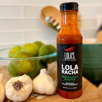 A bottle of Lola's Sriracha Sauce "Lola Racha" next to garlic and limes, perfect for grilling, dipping, or drizzling.