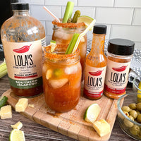 A group of Lola's Original Hot Sauce bottles on a cutting board, featuring a jar of hot sauce, citrus-infused liquid with limes, and a bowl of green olives.