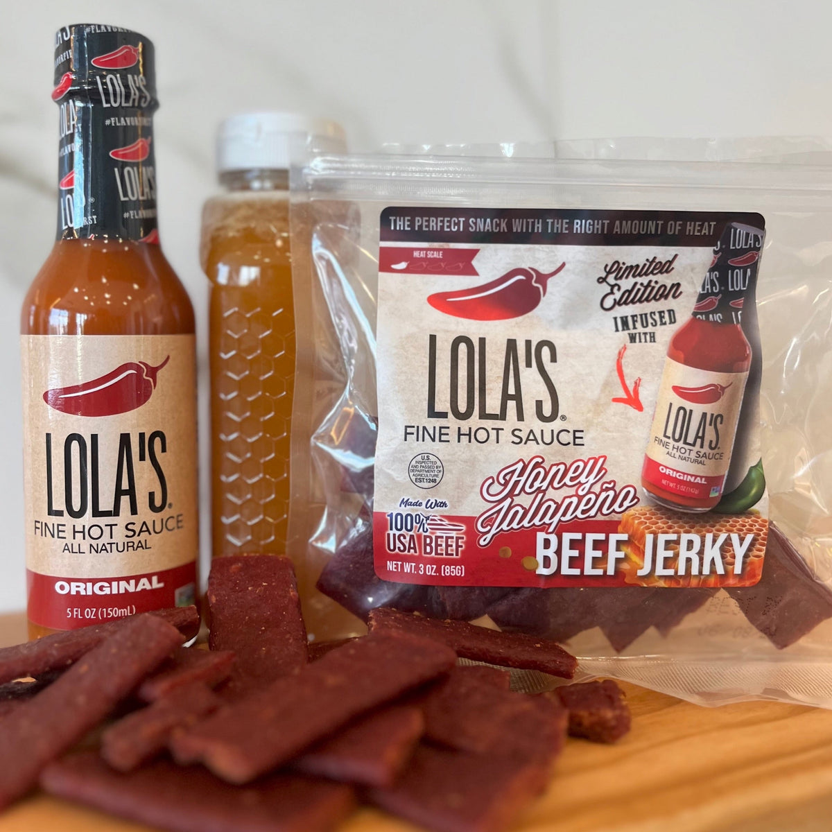 A group of food items on a table, including a bottle and package of Lola's Fine Hot Sauce's Honey Jalapeño Beef Jerky.